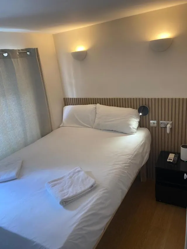 A double bed with white linen and two folded towels sits beside a wood-slatted headboard wall, with a black bedside table and a curtained window on the left.