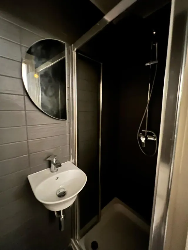A small bathroom with a wall-mounted white basin, a round mirror fogged with steam, and a glass-enclosed shower with a handheld head on a wall bar.