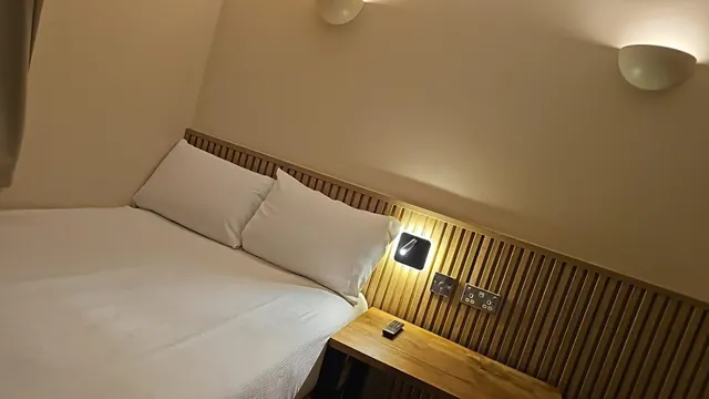 A double bed with white pillows sits beside a slatted wooden headboard wall, with a wall-mounted reading light and a remote control on a wooden bedside table.