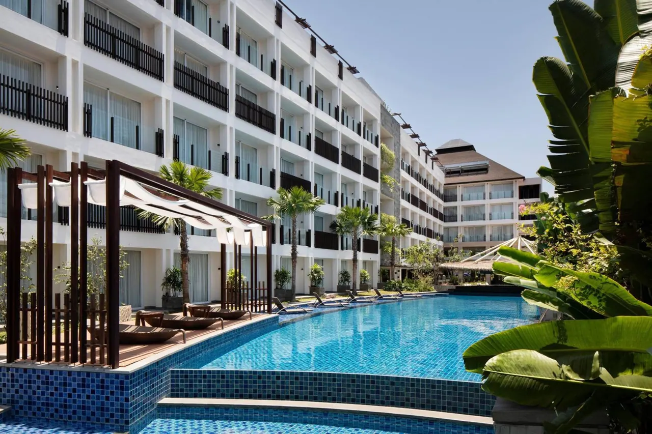 A large blue swimming pool with sun loungers lies beside a multi-storey white hotel building with balconies, with palms and banana plants along the edge.