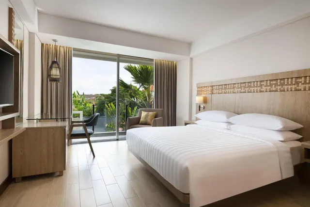 A king bed with white striped linen faces a large glass door onto tropical greenery, with an armchair, a desk and a wall-mounted television in the room.