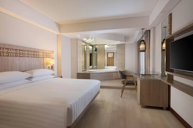 A king bed with striped white linen stands beside a raised bathtub, a glass-topped desk and a wall-mounted television in a hotel bedroom.