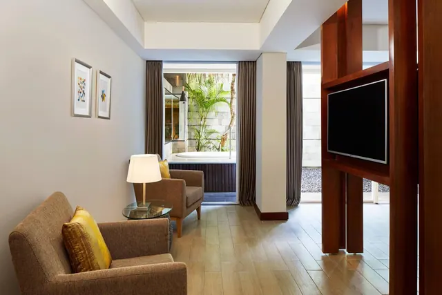 A living area with two armchairs, a side table with a lamp and a wall-mounted television, with glass doors looking out to a courtyard containing a small plunge pool and palm trees.