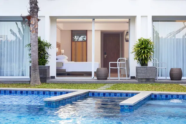 A white villa with open sliding glass doors shows a bedroom with a double bed, beside a tiled pool and potted plants.