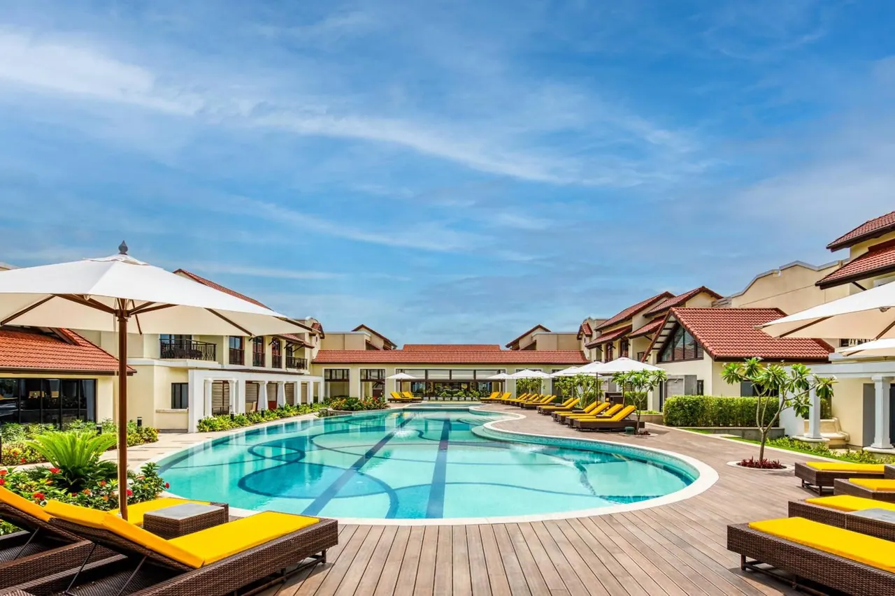A large outdoor pool is surrounded by sun loungers, umbrellas, landscaped gardens and tiled resort buildings.