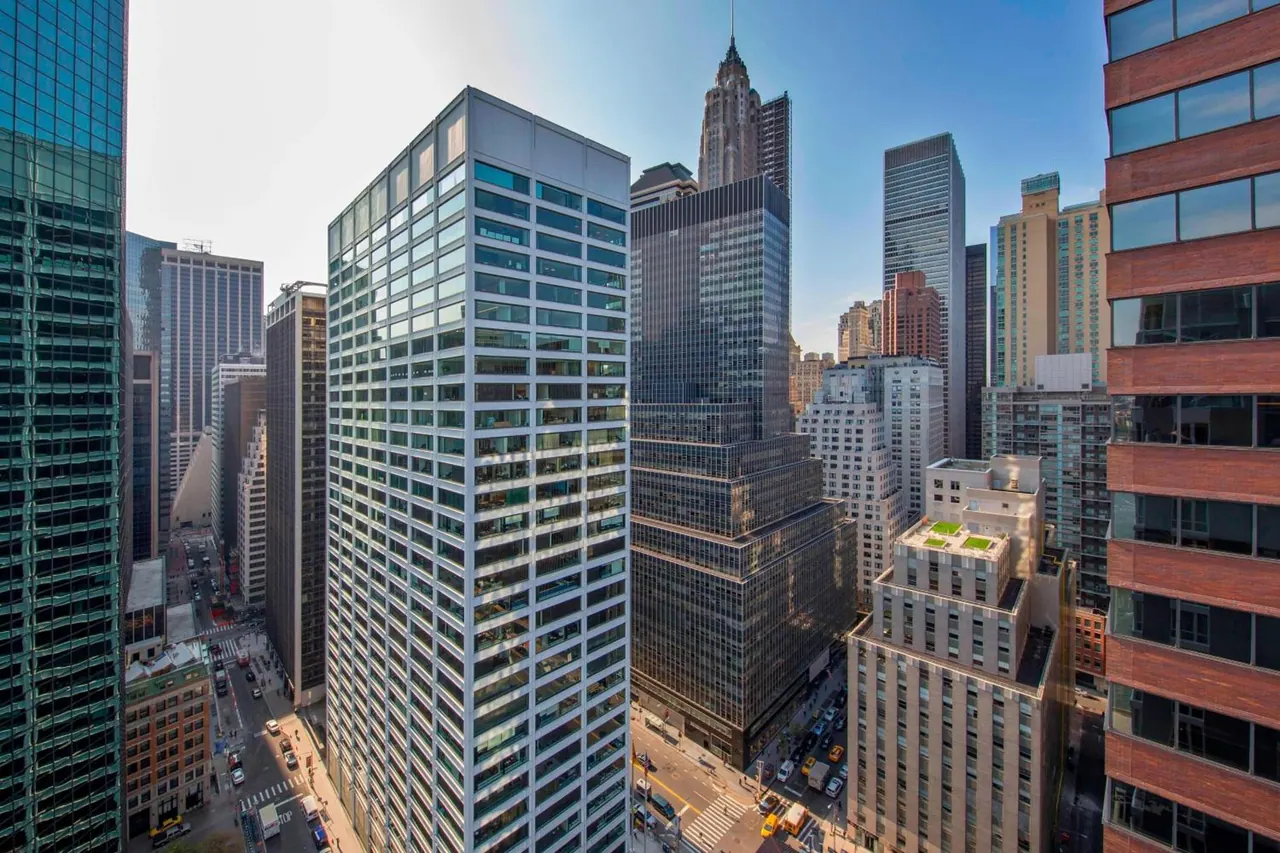 A high view looks down past glass and stone skyscrapers onto Midtown streets, with the stepped crown of the Chrysler Building rising in the background.