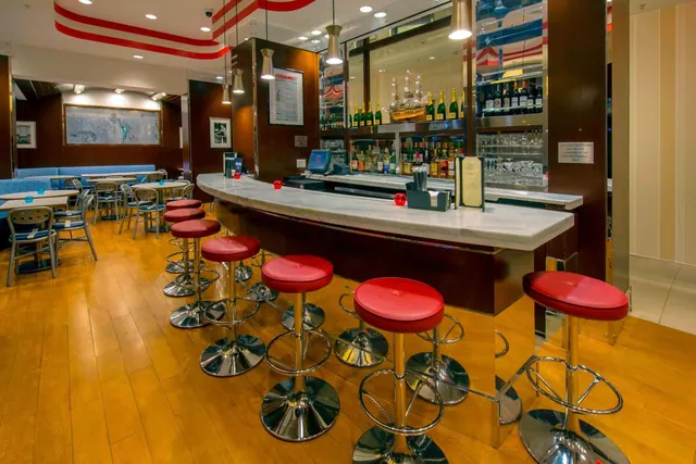 A curved marble bar with red leather stools, backed by shelves of bottles and glassware, with blue booth seating and tables in the background.