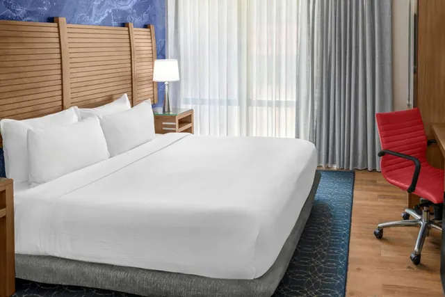 A king bed with white bedding and a wooden slatted headboard sits beside a desk with a red chair in a hotel bedroom.