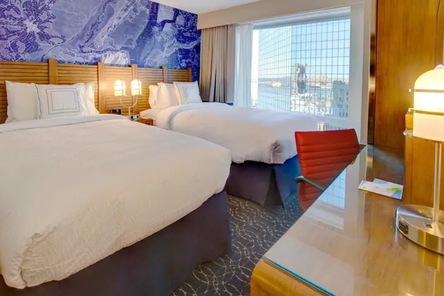 A hotel room with two double beds, a blue map-print wall, a red leather desk chair, and a window looking out over buildings and water.