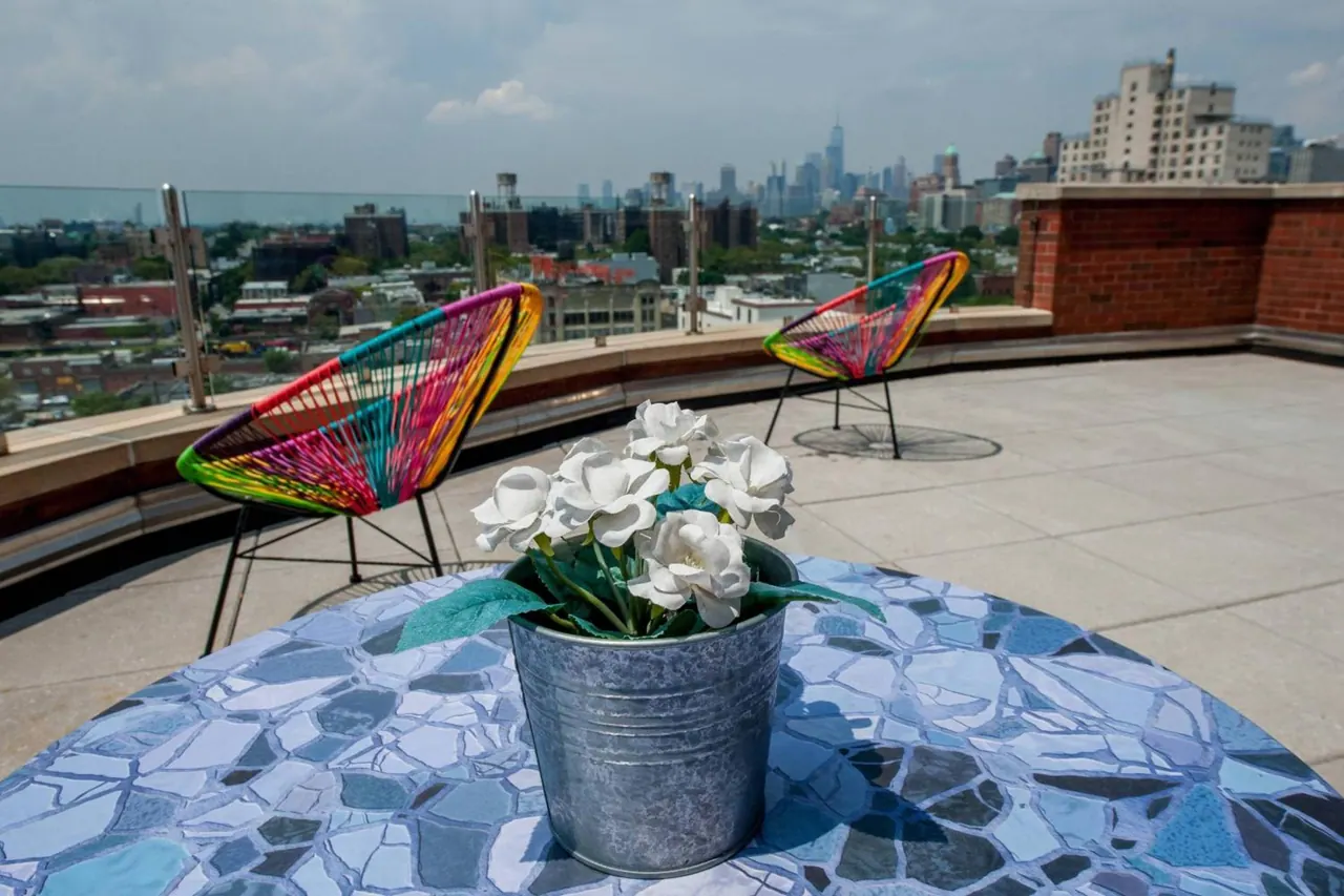 A rooftop terrace with colorful woven chairs and a mosaic table holding a metal bucket of white flowers, with the Manhattan skyline visible in the background.