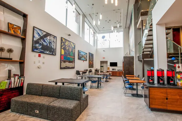 A lobby with a grey modular sofa, tables and chairs, framed artwork on the walls, and red coffee dispensers on a wooden counter.