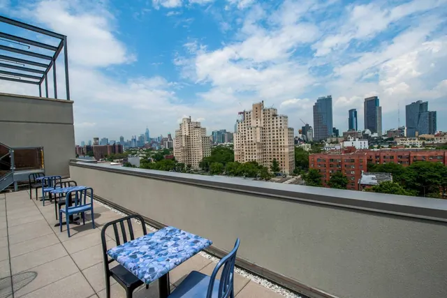 A rooftop terrace with blue metal tables and chairs overlooks a city skyline of high-rise towers and tree-lined streets under a partly cloudy sky.