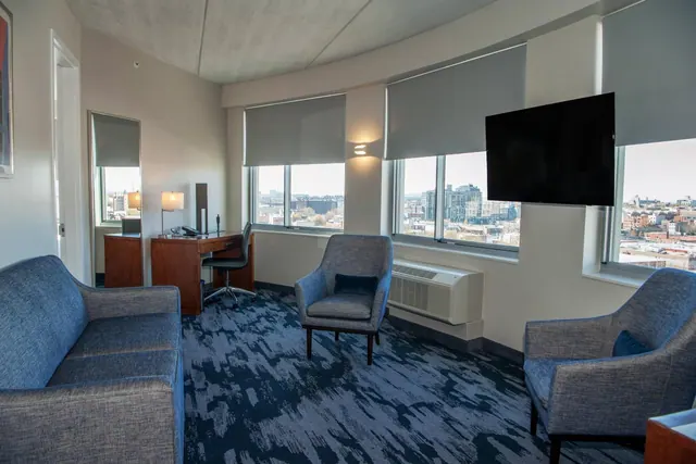 A seating area in a hotel room with a blue sofa, two armchairs, a wall-mounted television, a desk, and windows overlooking a city.