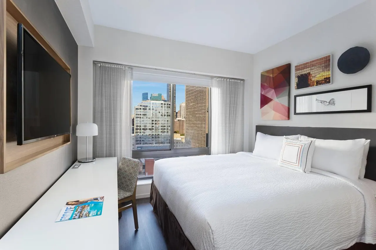 A bedroom with a king bed, a wall-mounted television, a desk with a chair, and a window showing city high-rises.