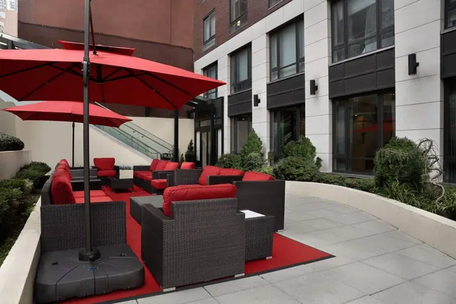 A paved outdoor terrace with red wicker sofas, red cushions, red patio umbrellas and a red carpet, beside a white stone building with large windows.