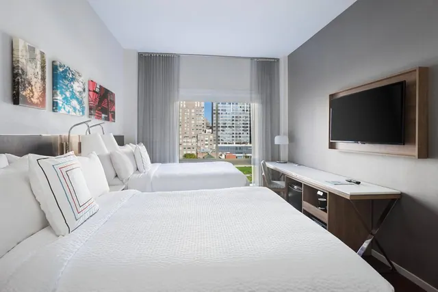 A hotel room with two white double beds, a wall-mounted flat-screen television, a desk with a small fridge beneath it, and a view of city buildings through the window.
