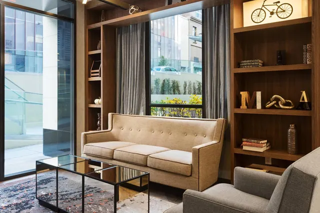 A lounge seating area with a tan sofa, grey armchair, glass coffee table and wood shelving, with a window looking onto a courtyard with plants and a building.