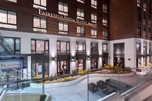 Exterior of a brick Fairfield Inn & Suites Marriott building at dusk, with an illuminated sign, landscaped planting, and an outdoor seating area behind glass railings.