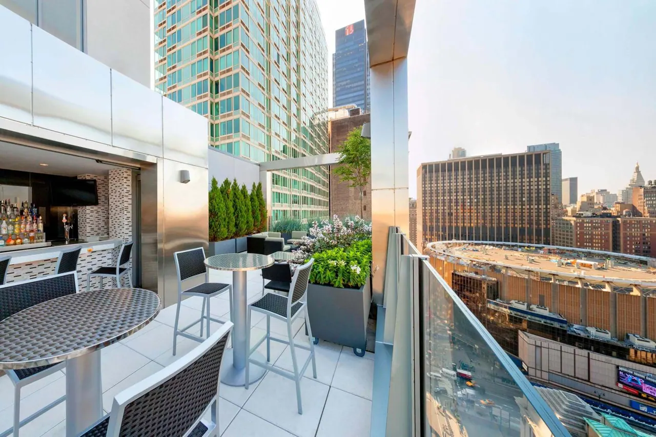 A rooftop terrace with a bar counter, high tables and stools, planters, and city buildings visible beyond the glass railing.