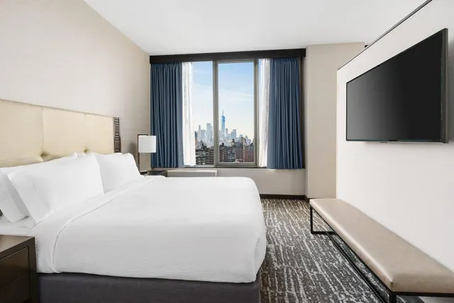 A bedroom with a king bed, a wall-mounted television and a bench at the foot of the bed, with a window showing a view of the Lower Manhattan skyline including One World Trade Center.