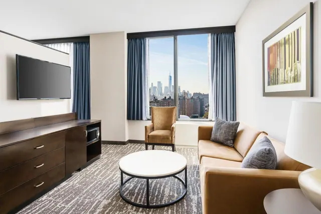 A living area with a wall-mounted television, a wood dresser, a tan sofa, an armchair and a round coffee table, with a window showing a city skyline that includes One World Trade Center.