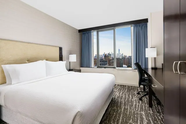 A king bed with white bedding sits across from a large window with a city skyline view, beside a dark wood desk and chair.