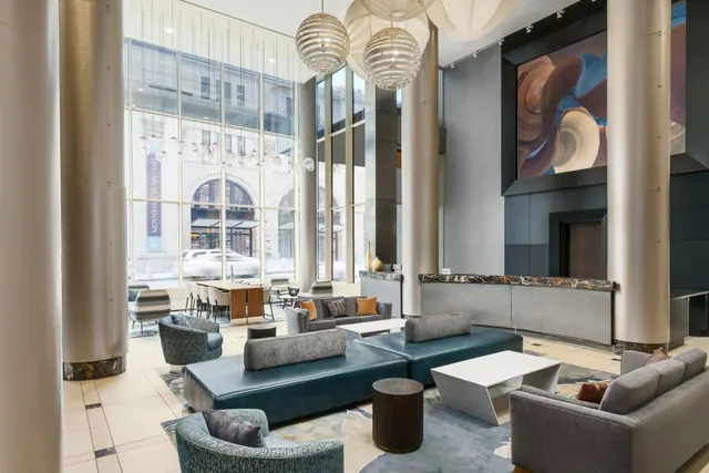 A hotel lobby with grey and teal sofas, white coffee tables, and tall windows looking onto a street and a historic building with a Moynihan Train Hall sign.