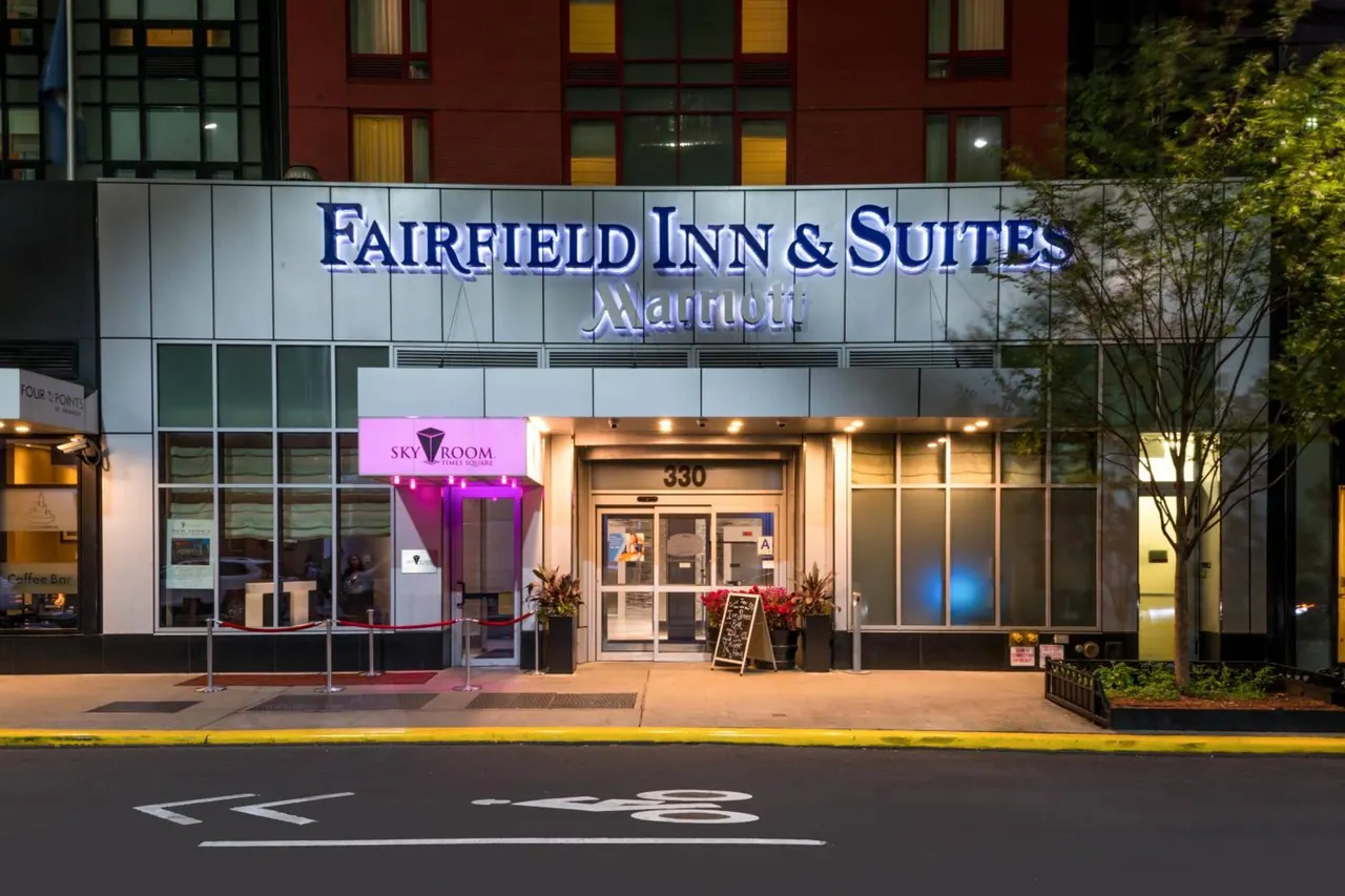 Street-level exterior of the Fairfield Inn & Suites Marriott at dusk, with the illuminated sign above the glass entrance doors and a small sandwich board on the pavement.
