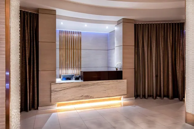 A reception desk with a marble front and a lit wood slat backdrop stands in a lobby with brown floor-length curtains and a tiled floor.