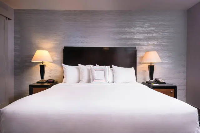 A king bed with white linens and two lit bedside lamps stands against a textured silver wall.