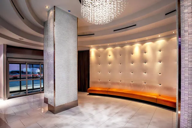 A hotel lobby with a curved orange leather bench against a tufted white wall, a crystal chandelier overhead, and glass entrance doors showing a street outside.