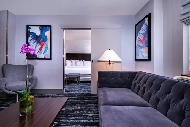 A living area with a tufted grey sofa, a grey armchair, a coffee table with an orchid, and a view through a sliding door to a bed with white linens.