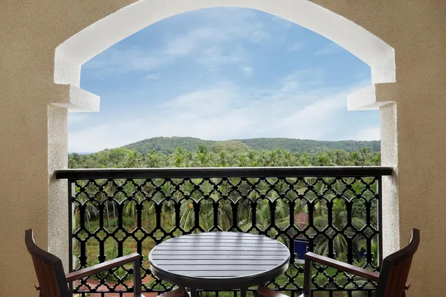 A furnished balcony overlooks palm trees and a green hillside.