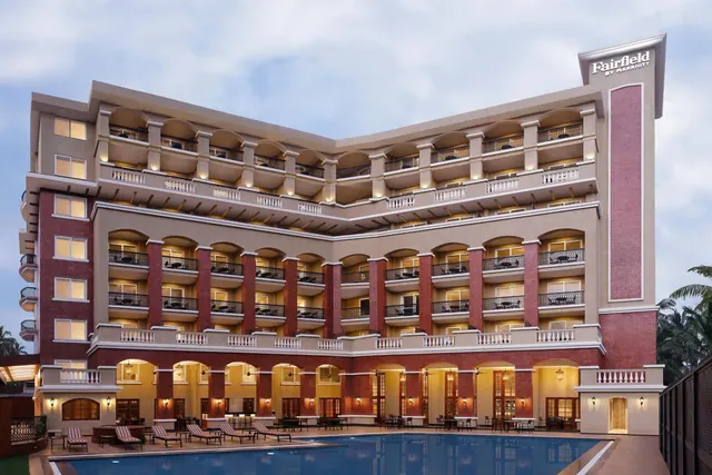 A multi-storey hotel building with arched balconies stands behind a large swimming pool and poolside loungers.