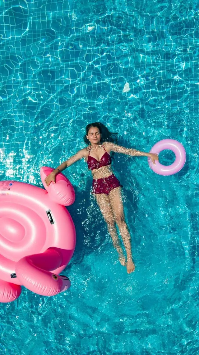A person floats in a bright blue swimming pool beside pink inflatable floats.
