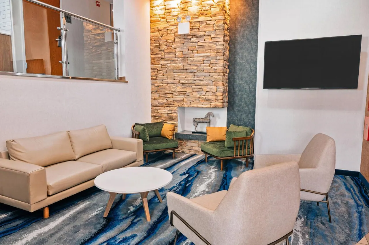 A lobby seating area with a beige sofa, upholstered armchairs, a round white coffee table, a stone accent wall with a fireplace, and a wall-mounted television.