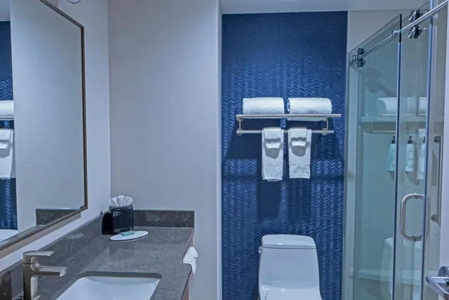 A bathroom with a single sink vanity, a toilet, folded towels on a rack against a blue tiled wall, and a glass-enclosed shower.