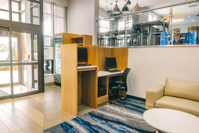 A lobby corner with two computer workstations and a black office chair, a beige sofa and a round white table, with glass entrance doors on the left.