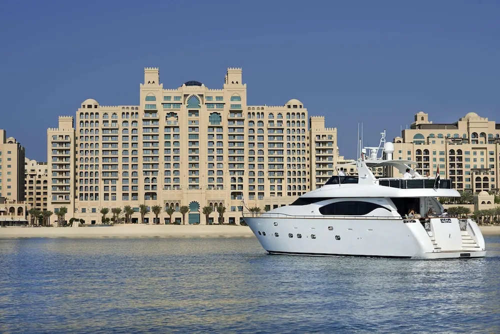 A large cream-coloured hotel building rises behind a sandy beach, with a white motor yacht on the water in the foreground.
