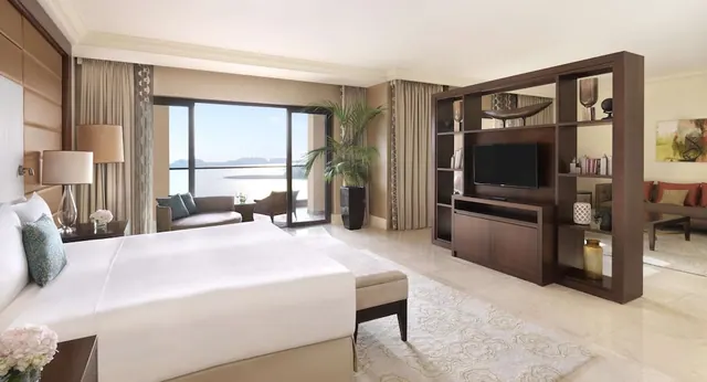 A hotel bedroom with a king bed, a seating area, a wall-mounted television and an open balcony door looking out over the sea.