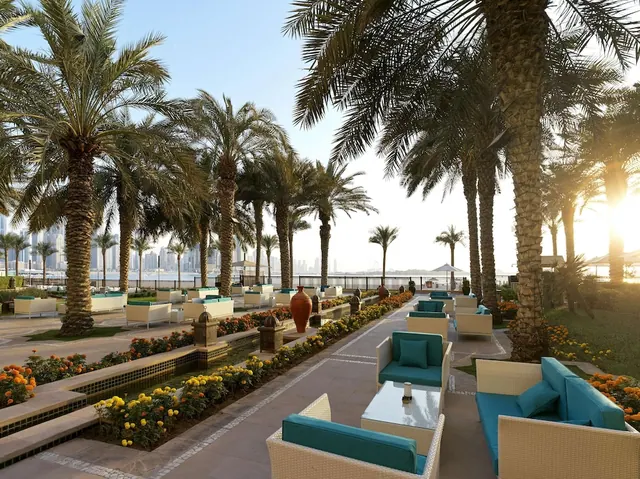 An outdoor terrace lined with palm trees, teal-cushioned lounge seating and a flowerbed beside a water channel, with a city skyline visible across the water.