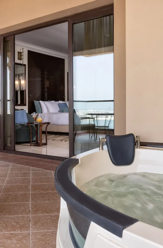 A jacuzzi sits on a balcony in the foreground, with sliding glass doors open to a bedroom containing a bed and a sofa and a pale sea view beyond.