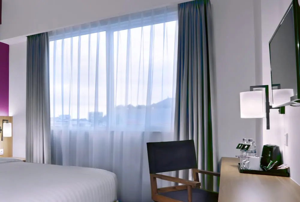 A hotel bedroom with a made bed, a sheer-curtained window showing hazy distant hills, a desk with water bottles, and a wall-mounted television.