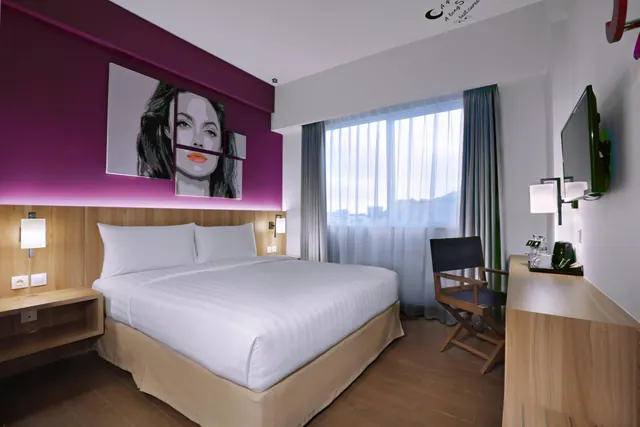 A bedroom with a king bed, a pop-art portrait on a magenta wall, a wall-mounted television and a wooden desk beside a large window.