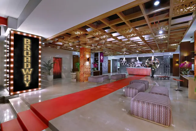 A hotel lobby with a red carpet runway, an illuminated Broadway-style sign, upholstered ottoman seating and a reception desk with a film-themed star logo.