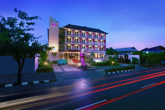 Exterior of a pink multi-storey hotel building lit up at dusk, with a minibus parked at the entrance and light trails from traffic on the road in front.