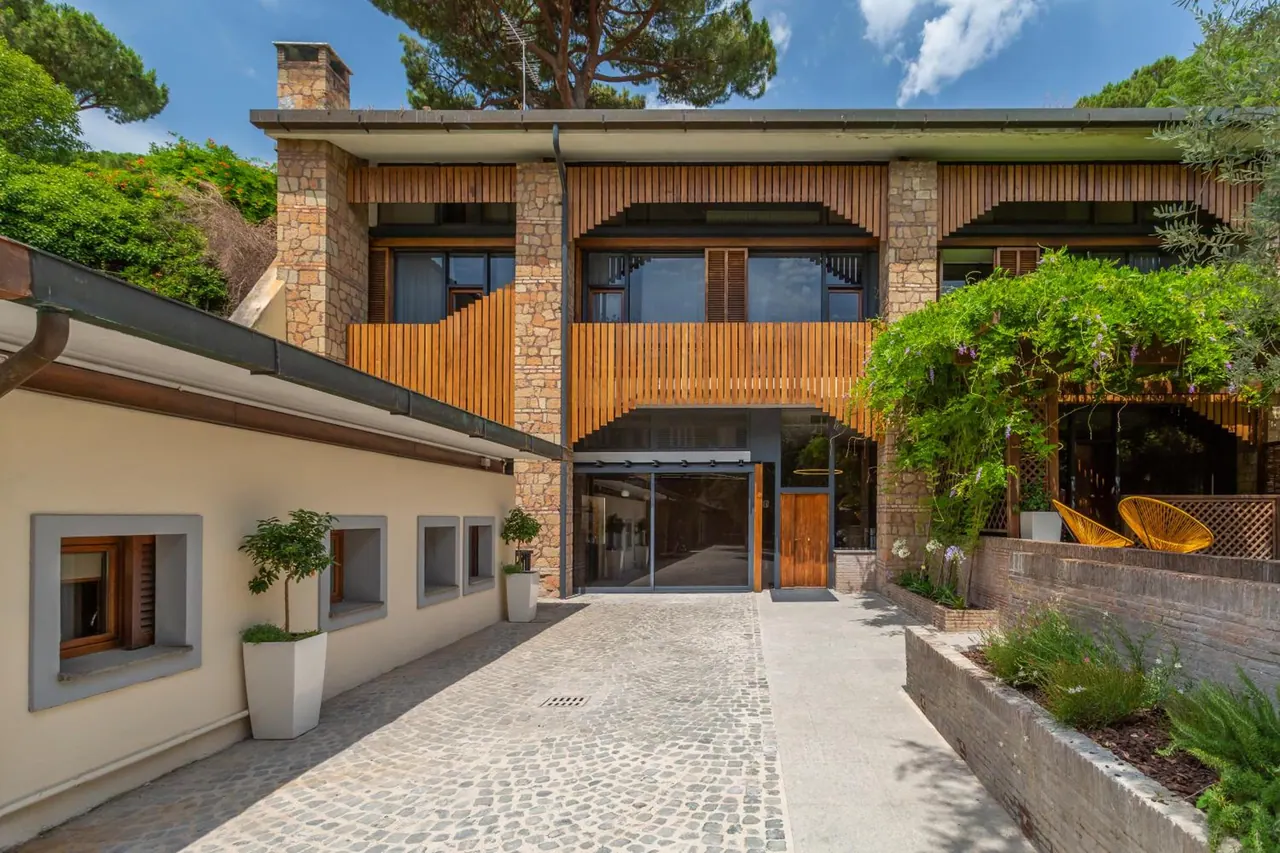 The front of a two-storey stone and timber building with a cobbled driveway, glass entrance doors and a wisteria-covered pergola beside a terrace with yellow chairs.