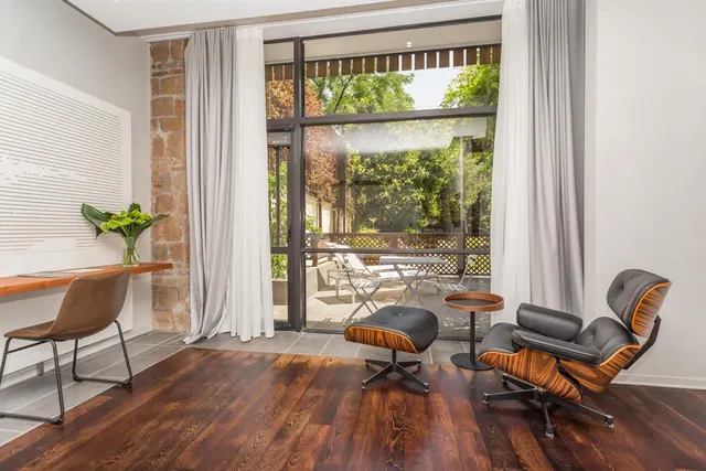A leather lounge chair with an ottoman and a small round side table sit on a dark wood floor beside floor-to-ceiling glass doors that open onto a terrace with chairs and green trees.