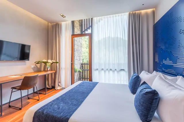 A bedroom with a double bed with blue accent pillows, a wall-mounted television, a wooden desk and chair, and a glass door opening onto a balcony with greenery beyond.