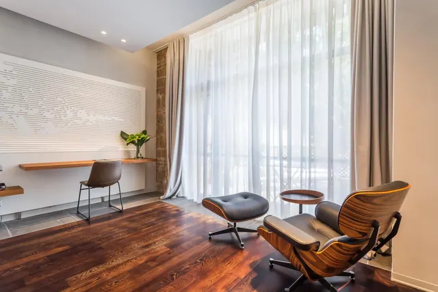 A leather lounge chair and ottoman sit on a wooden floor beside a large window with sheer curtains, with a wall-mounted desk and chair on the left.